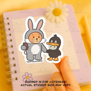 May include: A sticker featuring a cartoon bunny in a gray jumpsuit holding a phone, and a black duck wearing a face mask. The text "ZOOMED IN FOR CUTENESS! ACTUAL STICKER SIZE MAY VARY." is at the bottom. The sticker is held by a hand.
