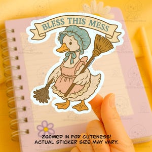 May include: A sticker featuring a cartoon goose wearing a bonnet and apron, holding a broom. The banner above the goose reads "BLESS THIS MESS." The sticker is on a spiral notebook. The text "ZOOMED IN FOR CUTENESS! ACTUAL STICKER SIZE MAY VARY." is at the bottom.
