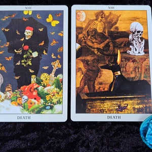 Puede incluir: Dos cartas de tarot que representan la carta de la Muerte. La carta de la izquierda presenta una figura esquel&eacute;tica con una capa negra que sostiene una rosa y un paraguas, rodeada de mariposas y flores. La carta de la derecha muestra una figura esquel&eacute;tica en una tumba con una estatua negra de Anubis. Ambas cartas tienen el n&uacute;mero romano XIII y la palabra "MUERTE" impresa en ellas.