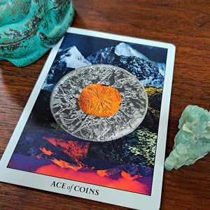 Puede incluir: Una carta de tarot con fondo blanco y una imagen colorida de una moneda con una escena de monta&ntilde;a. La carta dice "ACE of COINS".