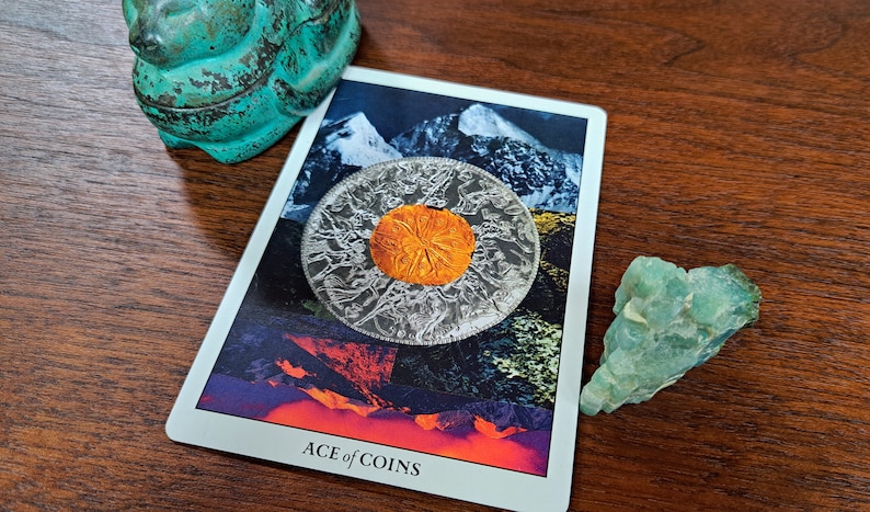 Puede incluir: Una carta de tarot con fondo blanco y una imagen colorida de una moneda con una escena de monta&ntilde;a. La carta dice "ACE of COINS".