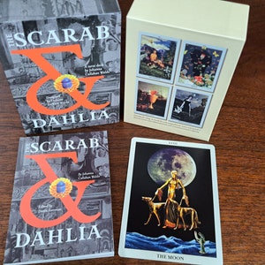 Puede incluir: Dos cajas con el texto "The Scarab & Dahlia" en la parte delantera. Una carta de tarot con el texto "The Moon" y una imagen de una mujer con dos perros frente a una gran luna.