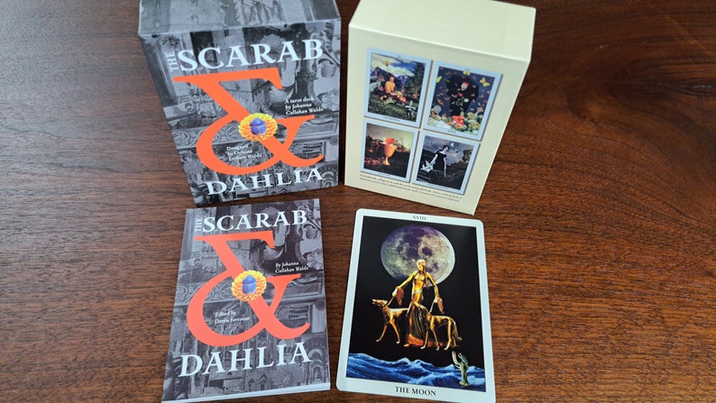 Puede incluir: Dos cajas con el texto "The Scarab & Dahlia" en la parte delantera. Una carta de tarot con el texto "The Moon" y una imagen de una mujer con dos perros frente a una gran luna.