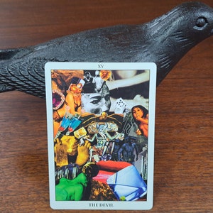 Puede incluir: Una carta de tarot con el n&uacute;mero XV y las palabras "THE DEVIL" impresas en la parte inferior. La carta presenta un collage de im&aacute;genes, que incluyen la cara de una mujer, el cuerpo de una mujer, la mano de un hombre y un par de dados.