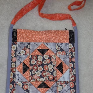 以下が含まれることがあります： グレー、ブラック、オレンジ、ホワイトの幾何学模様の生地で作られたパッチワークのハンドバッグ。バッグには長いオレンジ色のストラップが付いています。
