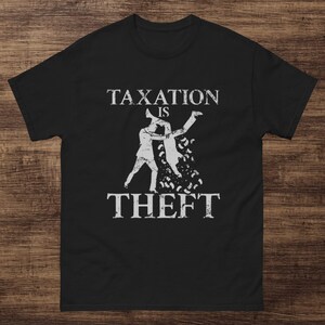 May include: Black t-shirt with a white graphic. The graphic depicts two figures, one holding the other upside down, with the words "TAXATION IS THEFT" in a bold font.