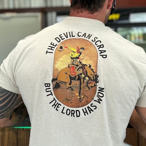 Puede incluir: Camiseta de color beige claro con un gráfico en la espalda que muestra a un vaquero montando un caballo salvaje. El texto dice "THE DEVIL CAN SCRAP BUT THE LORD HAS WON". También se ve una gorra de camionero negra y gris.