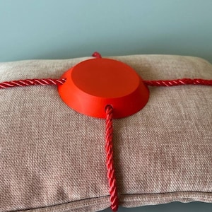 May include: A red circular pillow with a red rope tied around it in a criss-cross pattern.