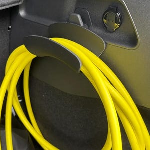 Op de afbeelding: Een close-up van het interieur van een auto toont een zwart opbergvak met een felgele oplaadkabel. De kabel is opgerold en wordt op zijn plaats gehouden door een zwarte houder. Een 12 V stopcontact is ook zichtbaar.