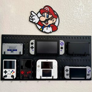 Könnte beinhalten: Eine schwarze Lochwand zur Wandmontage mit acht tragbaren Spielkonsolen, darunter eine graue Super DS Lite, eine weiß-graue Switch, ein schwarzer 3DS, ein weißes Retro-Gerät, eine transparente schwarze Konsole, ein weißer 2DS und ein graues Handheld.