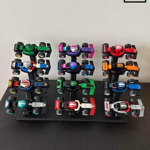 Peut inclure: Une collection de voitures de course miniatures de différentes couleurs, dont le vert, le violet, le rouge et l'orange, exposées sur des présentoirs noirs. Chaque voiture présente un casque de pilote et un design détaillé.