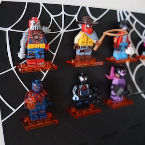 Può includere: Una bacheca nera con un design a ragnatela bianca, che contiene sette colorate minifigure Lego a tema supereroi. Le figure sono su supporti trasparenti arancioni. Le figure includono un robot, un cowboy e altri personaggi.