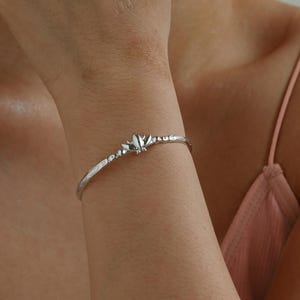 Könnte beinhalten: Ein silberfarbenes Armband mit einem strukturierten, organischen Design. Das Armband hat ein zentrales dekoratives Element und wird am Handgelenk getragen. Der Schmuck ist ein zartes Accessoire.
