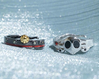 Könnte beinhalten: Zwei silberne Ringe mit schwarzen und goldenen Akzenten. Ein Ring hat einen roten Streifen und ein goldenes sternförmiges Design. Der andere Ring hat ein schwarzes Gesicht mit zwei runden Augen.