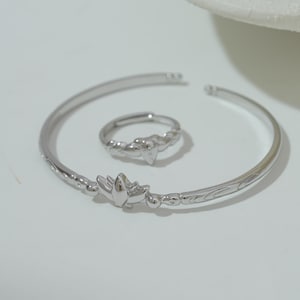 Könnte beinhalten: Ein silberfarbenes Schmuckset mit einem Armreif und einem Ring. Der Armreif hat ein florales Design und der Ring hat ein verdrehtes Band mit herzförmigem Akzent. Das Set wird auf einer weißen Oberfläche präsentiert.