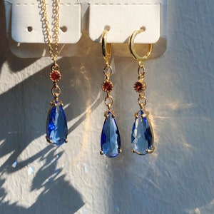 Op de afbeelding: Een set gouden oorbellen en een ketting met blauwe traanvormige kristallen en rode accenten.