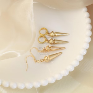 May include: Three pairs of gold earrings with a pointed design. Two pairs are hoop earrings with a pointed dangle, and one pair is a drop earring with a pointed dangle.