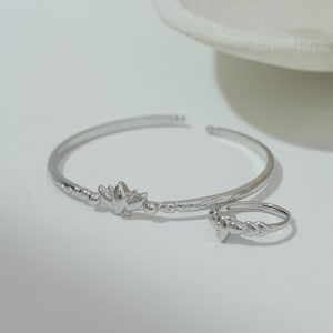 Könnte beinhalten: Ein silberfarbenes Armband- und Ring-Set. Das Armband hat ein Blumendesign in der Mitte und ist an den Enden offen. Der Ring hat ein geflochtenes Band und ein passendes Blumendesign. Der Schmuck wird auf einer weißen Oberfläche präsentiert.