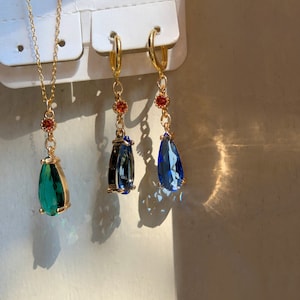 May include: Three gold teardrop earrings with blue and green gemstone accents. The earrings have a small red stone at the top of the teardrop.