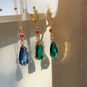 Op de afbeelding: Drie gouden kettingen met traanvormige hangers. Eén hanger is blauw, de andere twee zijn groen.