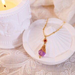 May include: A gold necklace with a purple and red glass pendant. The pendant is shaped like a teardrop and has a delicate filigree design. The necklace is on a white surface.