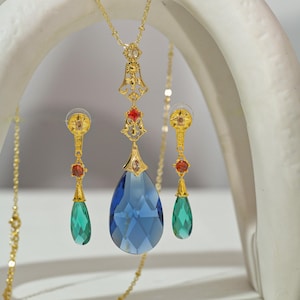 May include: A gold necklace and earrings set featuring a teardrop-shaped blue crystal pendant and matching earrings. The earrings have a green crystal drop and a red crystal accent.