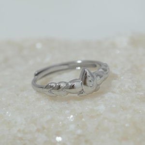 Könnte beinhalten: Ein silberfarbener Ring mit floralem Design. Der Ring hat ein geflochtenes Band und ein zentrales blütenförmiges Element. Der Ring ist vor einem weißen, strukturierten Hintergrund platziert.