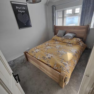 May include: A bedroom with a wooden bed frame and a yellow floral duvet. The bed is against a white wall with a black framed print that says "Here I Go Again" with a heart shape made of words. The room has grey carpet and a window with white blinds and grey curtains.