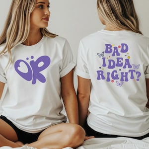 May include: White t-shirts with purple graphics. One shirt has a stylized "OR" design, the other says "BAD IDEA RIGHT?" in purple with butterflies, stars, and smiley faces. The shirts have rolled sleeves.