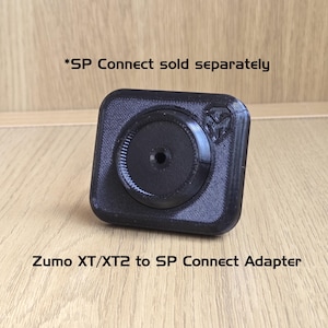 Pode incluir: Adaptador preto Zumo XT/XT2 para SP Connect. O adaptador é quadrado com um centro circular e uma superfície texturizada. O texto "*SP Connect sold separately" está acima.