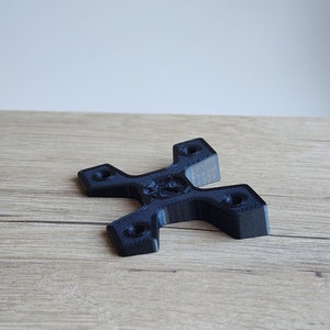 Puede incluir: Un soporte negro impreso en 3D con un diseño en forma de cruz. El soporte tiene cuatro agujeros circulares y una superficie texturizada. El objeto está sobre una superficie de madera.