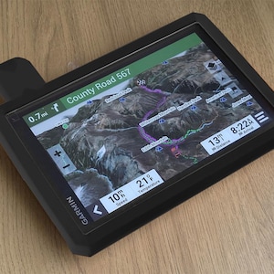 May include: A black Garmin GPS device displaying a detailed topographical map with a highlighted route. The screen shows "County Road 567", distance, speed, and temperature. The device is on a wooden surface.