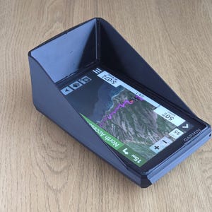 Pode incluir: Um dispositivo GPS Garmin preto com um ecrã que mostra um mapa e informações de rota. O dispositivo está montado num suporte preto inclinado. O ecrã mostra a hora, a velocidade e a distância. O dispositivo está numa superfície de madeira.