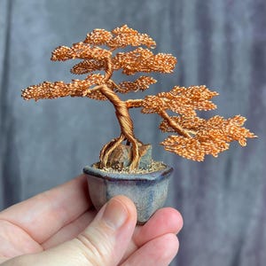 May include: A copper wire bonsai tree sculpture. The tree has a twisted trunk and detailed branches with textured leaves. It is planted in a small, square, blue ceramic pot. The sculpture is held in a person's hand.