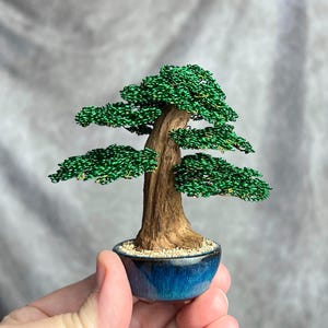 May include: A miniature bonsai tree sculpture featuring a textured brown trunk and vibrant green wire foliage arranged in layered branches. The tree is planted in a small, round, glossy blue ceramic pot filled with tiny light-colored pebbles.