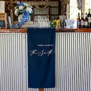 May include: A navy blue banner with the text "RAISE A GLASS TO The Smith" in white script. The banner is displayed on a bar with drinks, glassware, and floral arrangements. The bar has a corrugated metal front.
