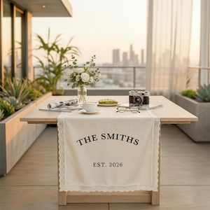 Könnte beinhalten: Ein Holztisch, der für eine Mahlzeit auf einer Dachterrasse gedeckt ist. Ein cremefarbener Tischläufer mit Spitzensaum zeigt "THE SMITHS EST. 2026". Eine Vintage-Kamera, eine Brille sowie eine Tasse und Untertasse befinden sich auf dem Tisch.