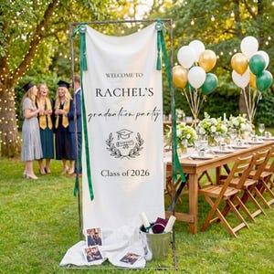 Puede incluir: Una pancarta blanca con detalles verdes da la bienvenida a la fiesta de graduación de Rachel. La pancarta, sostenida por un marco metálico, muestra el texto "Clase de 2026" y un gráfico de birrete y diploma. Globos y una mesa decorada en el fondo.