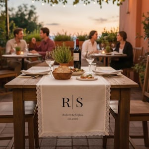 Puede incluir: Una mesa de madera rústica preparada para una cena, con un camino de mesa blanco con ribete de encaje y las iniciales "R S", los nombres "Robert & Sophia" y la fecha "est.2026". Una botella de vino tinto, pan y platos están sobre la mesa.