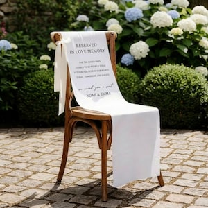 May include: A wooden chair with a white fabric draped over it, bearing the text "RESERVED IN LOVE MEMORY" and a message. The chair is set outdoors with a floral backdrop, ideal for a wedding or memorial event.