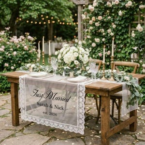 Może przedstawiać: Drewniany stół ustawiony na przyjęcie weselne, ozdobiony koronkowym bieżnikiem z napisem "Just Married", imionami i rokiem 2026. Stół jest udekorowany białymi kwiatami, świecami i zielenią.