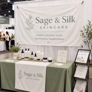 May include: A trade show booth display for Sage & Silk Skincare featuring a large white banner with green text, a sage green tablecloth with a branded runner, various skincare bottles and jars on a wooden tray, and a tiered brochure stand.