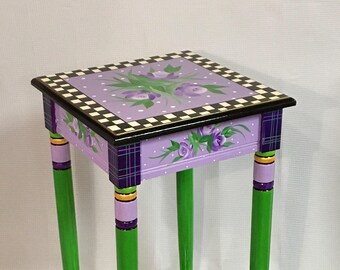 May include: A hand-painted side table with a checkerboard design on the bottom shelf and a purple top with a floral design and black and white checkerboard border. The legs are painted green with purple, orange, and black accents.
