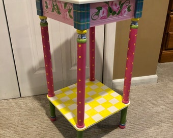 May include: A small, hand-painted side table with a pink, yellow, and green checkered design. The table has a square top and a lower shelf. The legs are painted with pink and yellow polka dots.