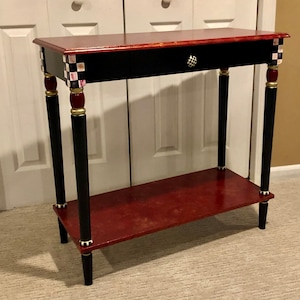 May include: A black and red console table with a single drawer and a lower shelf. The table has black painted legs with gold accents and a checkerboard pattern on the drawer front.