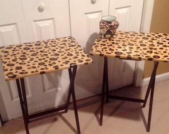 Puede incluir: Dos mesas plegables marrones con patas de metal negro. Las tapas de las mesas están cubiertas con un patrón de estampado de leopardo.