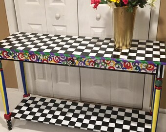 May include: A black and white checkered console table with a lower shelf. The table has a colorful painted design on the drawer and legs. The legs are painted yellow, blue, red, and green. A vase of flowers sits on top of the table.