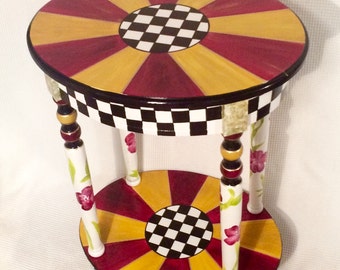 May include: A two-tiered round side table with painted floral details on the legs. The tabletops feature a black and white checkerboard design surrounded by alternating red and yellow stripes.
