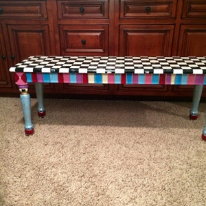 May include: A black and white chequered bench with a colourful striped border. The bench has blue, pink, and purple stripes with gold accents. The legs are painted blue with gold swirls and red accents.