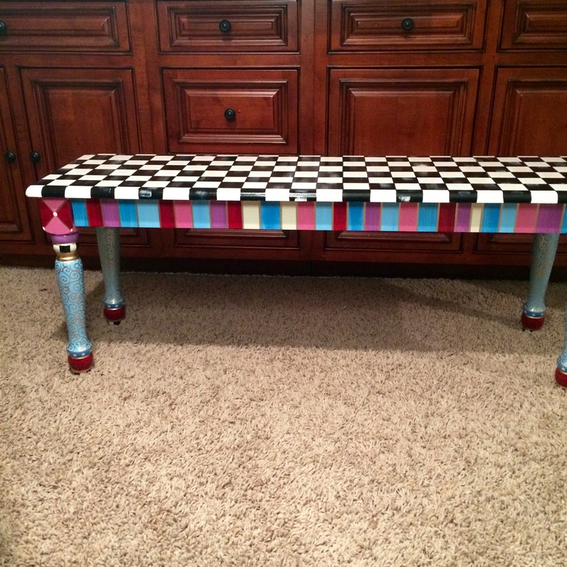 May include: A black and white chequered bench with a colourful striped border. The bench has blue, pink, and purple stripes with gold accents. The legs are painted blue with gold swirls and red accents.